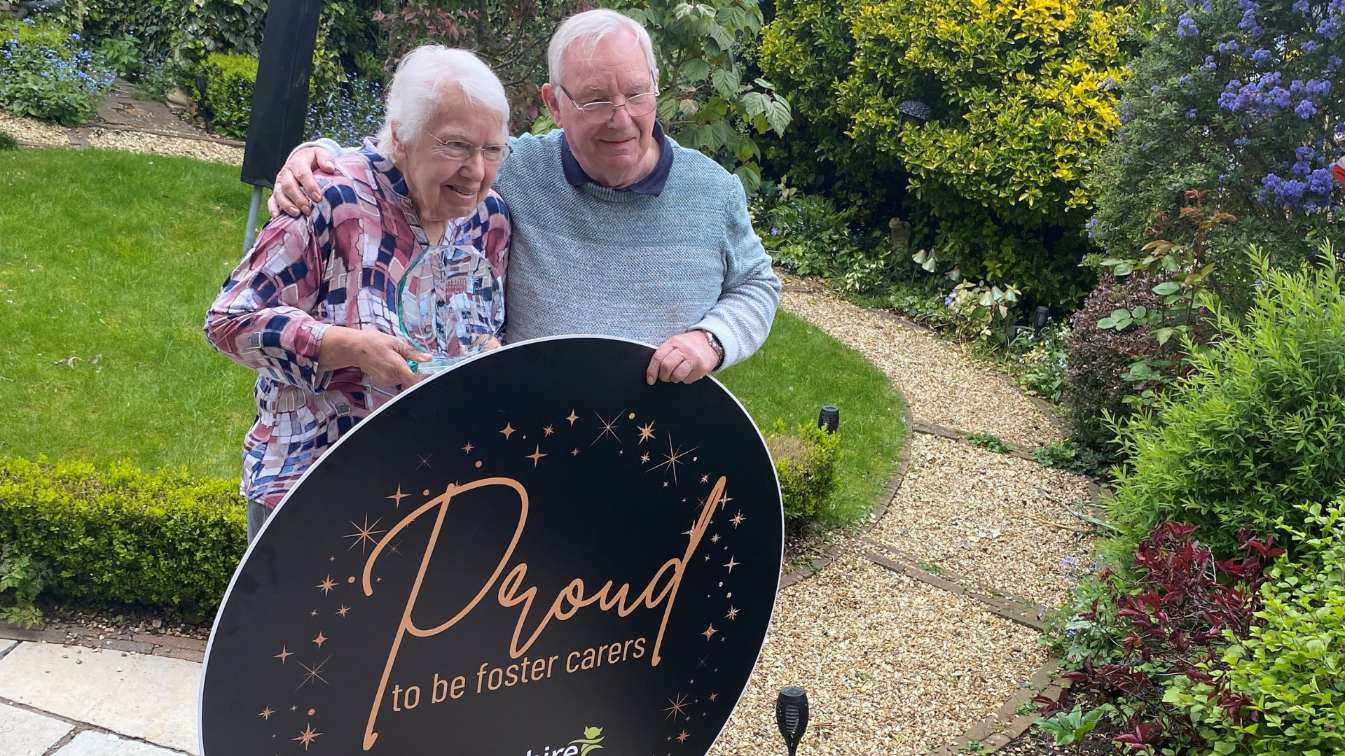 Rob and Marg stood holding a circular saying Proud Foster Carers in a garden setting
