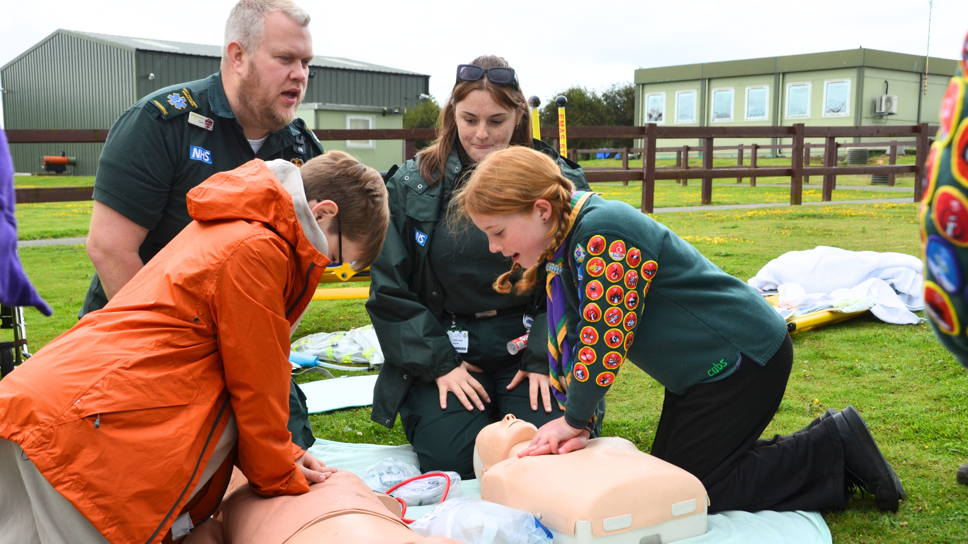 two children giving CPR to dummies, under instruction of two paramedics