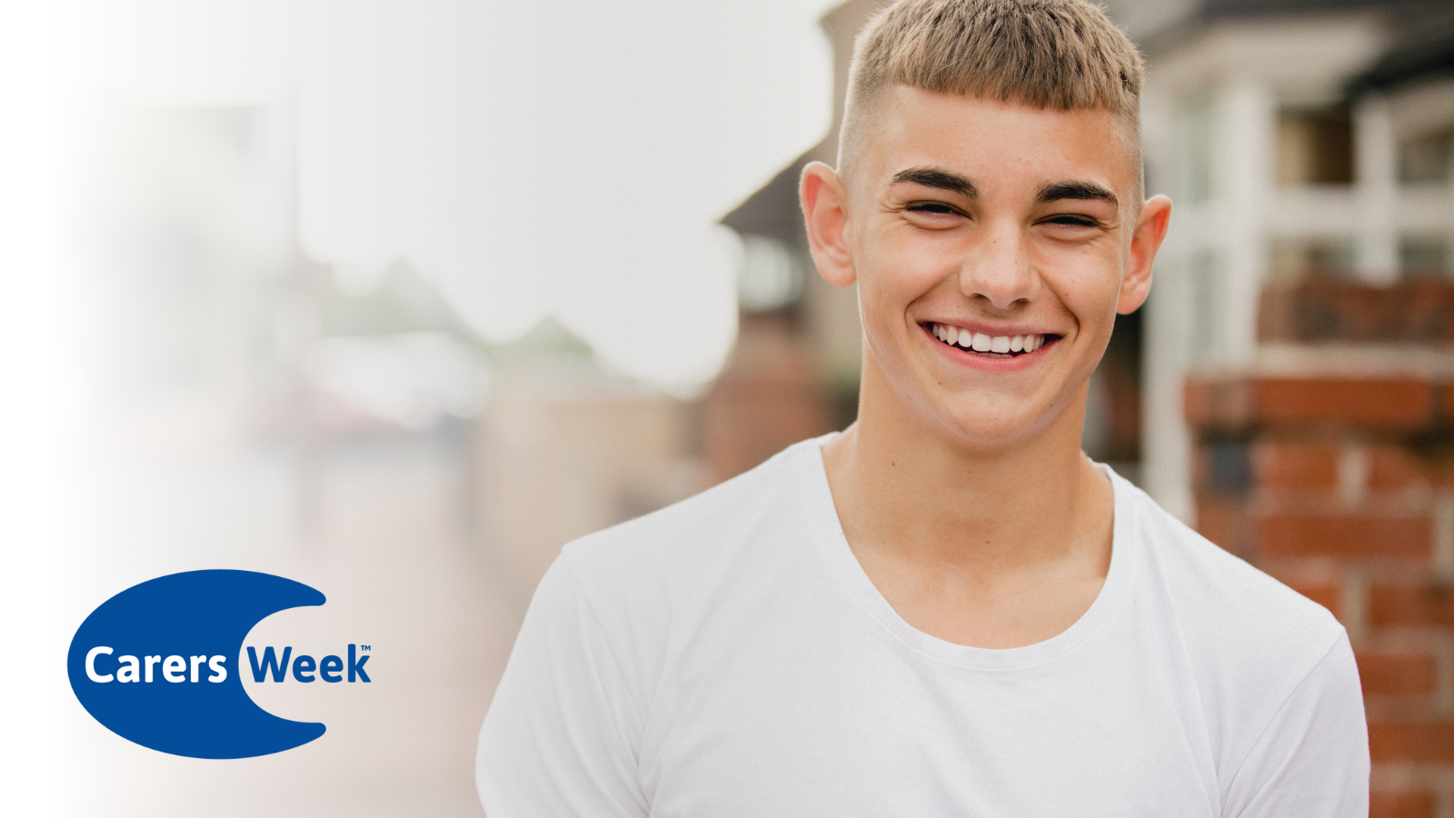 A young male adult wearing a white t-shirt smiling in the street with the carers week logo on.