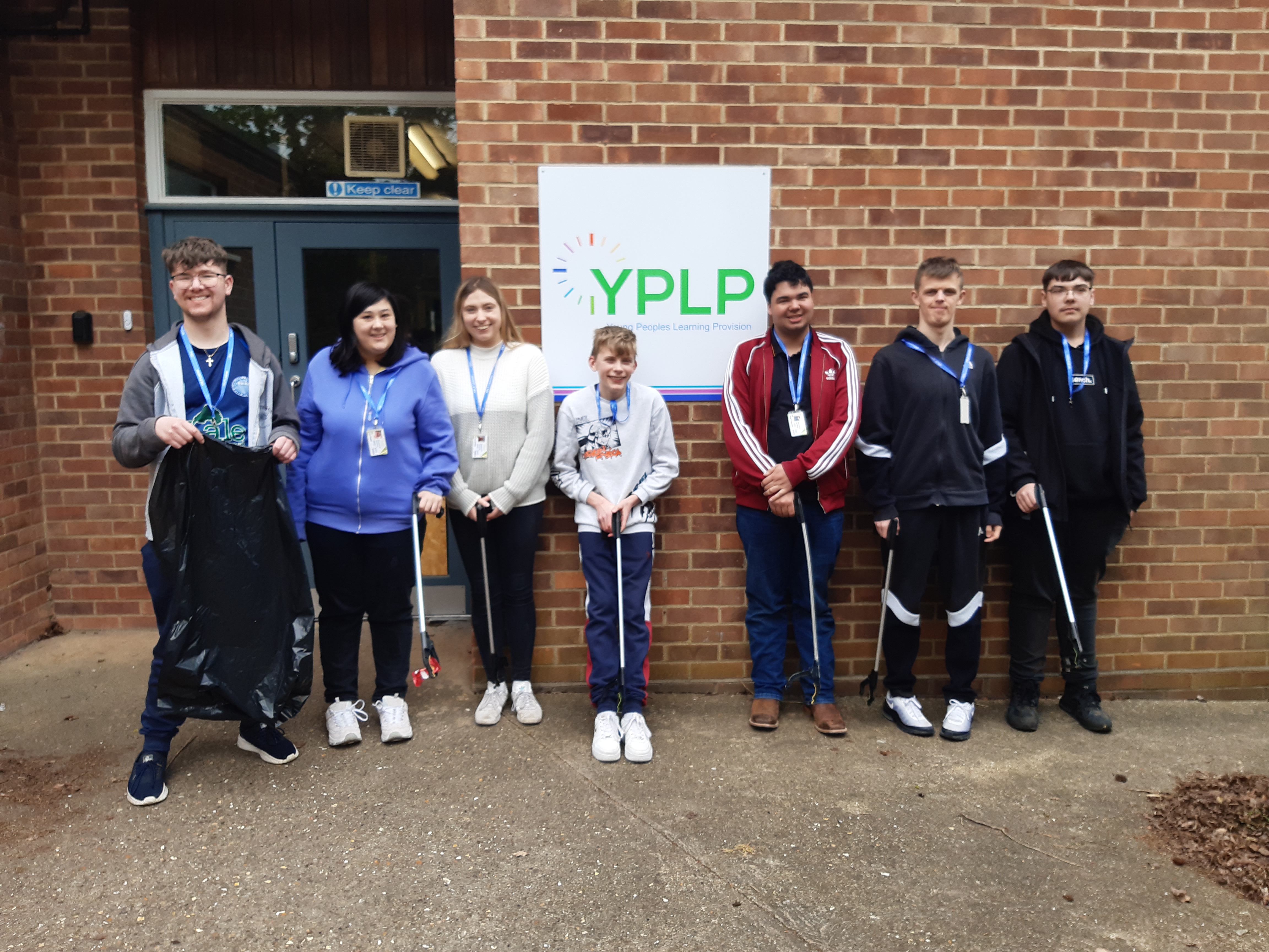 Young adults stood in a line outside a building, holding litter pickers and bin bags
