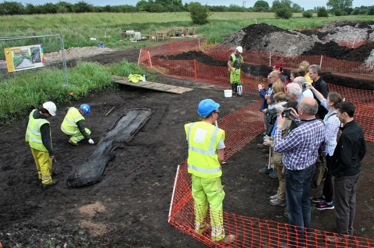 Archaeological excavation in progress, with workers uncovering a large preserved timber structure as members of the public watch from a fenced viewing area.