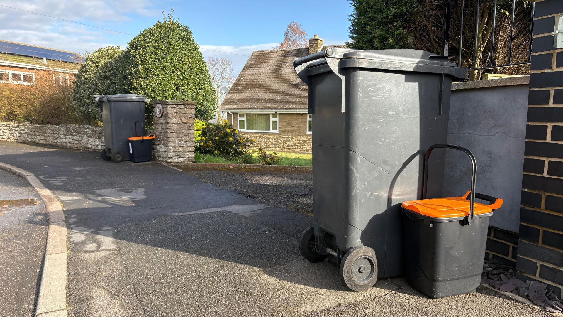 A grey wheeled waste bin and smaller grey food waste caddy with an orange lid presented together at the edge of a residential property. Another bin and food waste caddy can be seen further along the street.