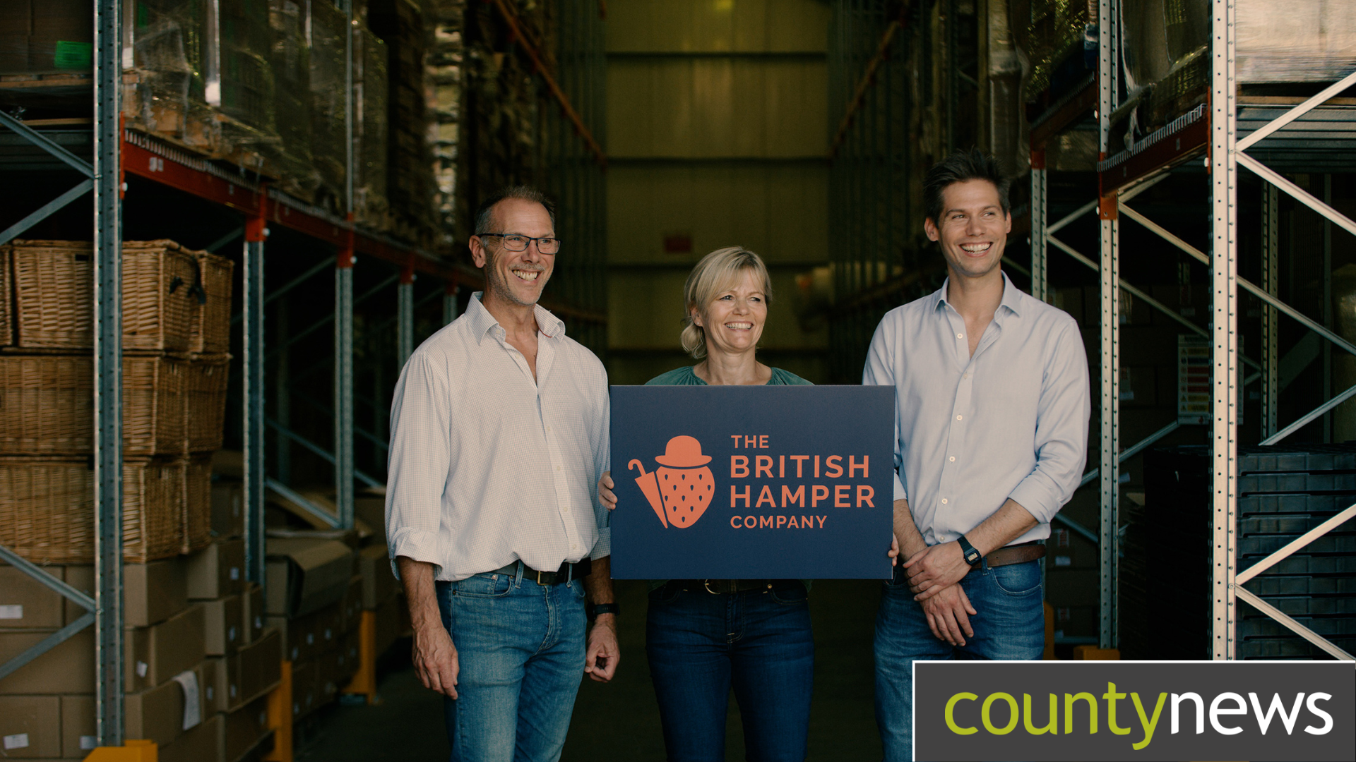 Man standing on the left, a woman in the middle holding up a sign with the text in orange saying The British Hamper Company, and a man to the right of her. Standing in a warehouse.