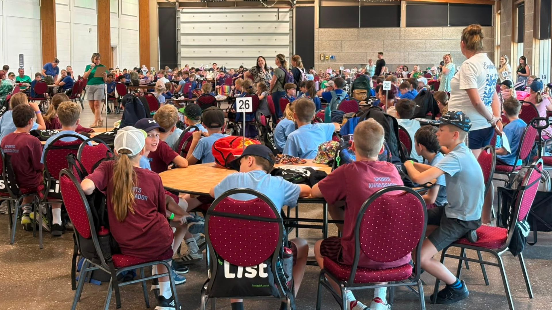 A large hall filled with hundreds of primary school pupils seated at numbered round tables during an educational event. Teachers and event staff stand among the groups, with one person speaking at the front of the room.