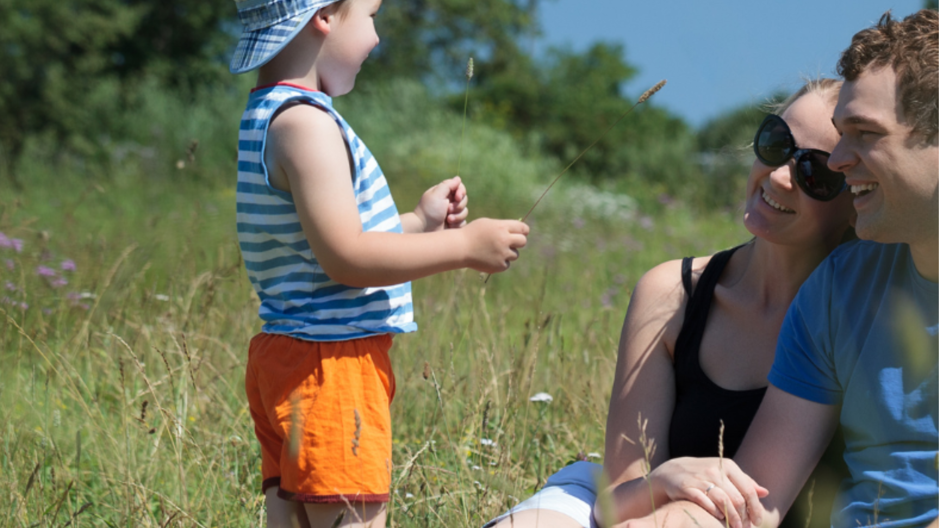 A family sat in a sunny field looking at their young son wearing a sun hat.