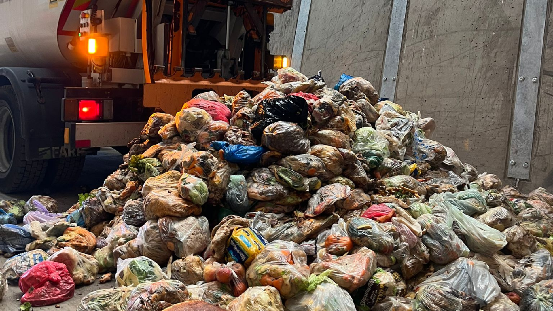 A large pile of bagged food waste sits on the ground behind a food waste truck, with plastic bags filled with discarded food waste stacked against a concrete wall.