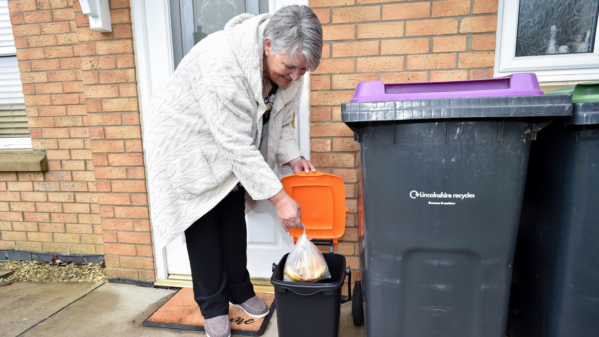 A woman outside a brick house places a tied liner containing food waste into a small black outdoor food waste caddy with an orange lid. The caddy is positioned beside larger household wheeled bins, including one labelled “Lincolnshire recycles.”
