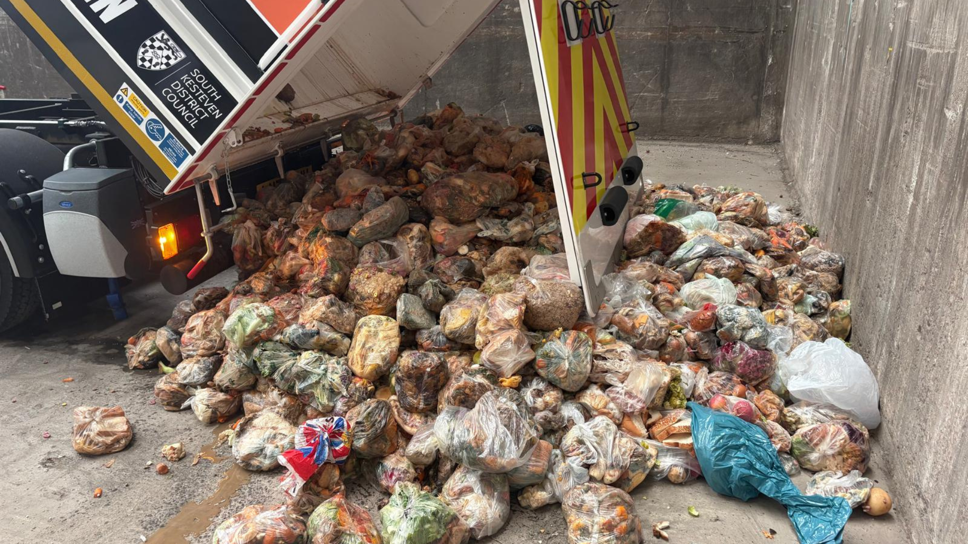 A food waste truck is tipping out a large pile of bagged food waste into a concrete waste area. The pile consists of numerous plastic bags filled with rotting food scraps, including vegetables and bread, some spilling onto the ground.