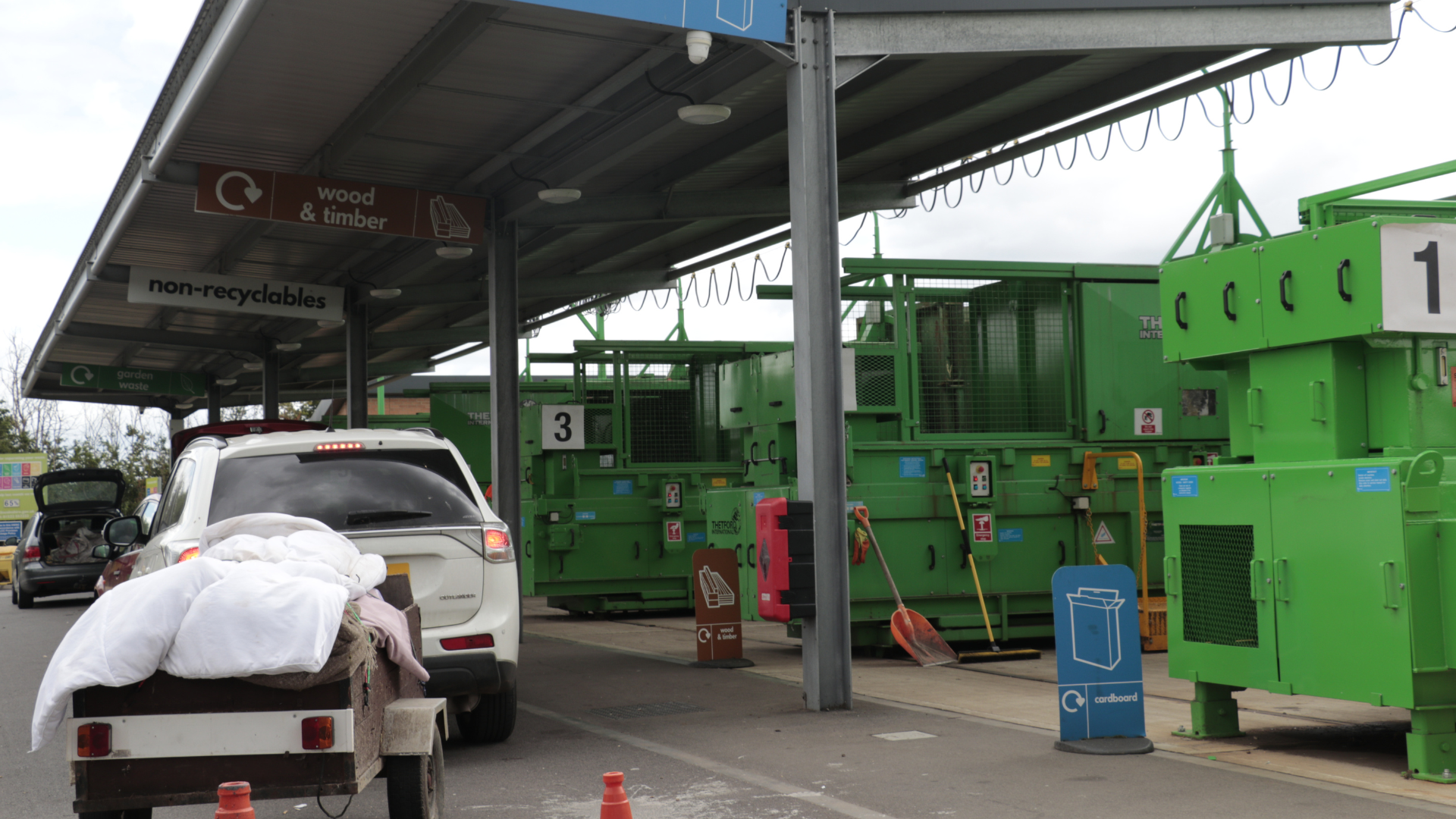 A vehicle towing a trailer loaded with mattresses and bedding waits at a household waste recycling centre, with clearly signed recycling bays and large green waste compactors visible under a covered area.