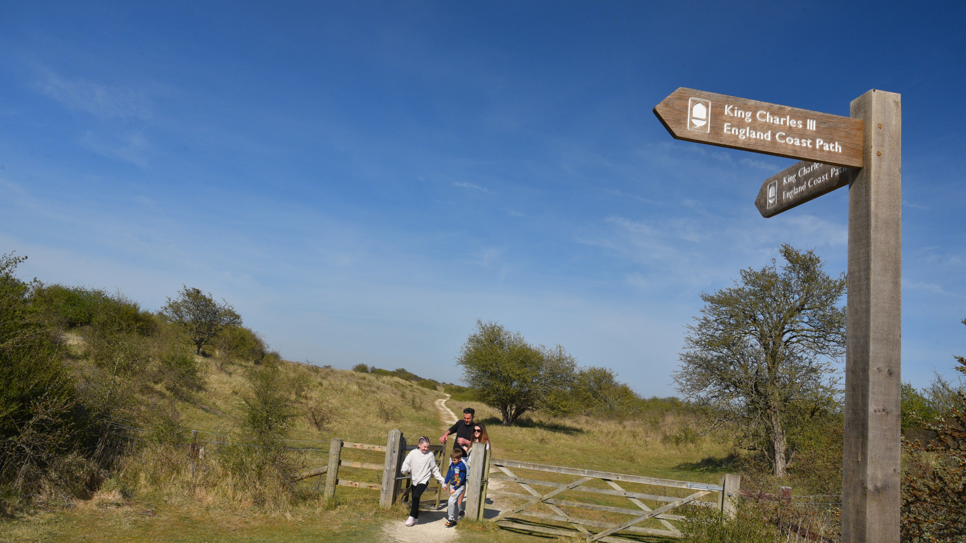 A family walking through a gate on a nature path with green grass and a sign post for the King Charles England coast path