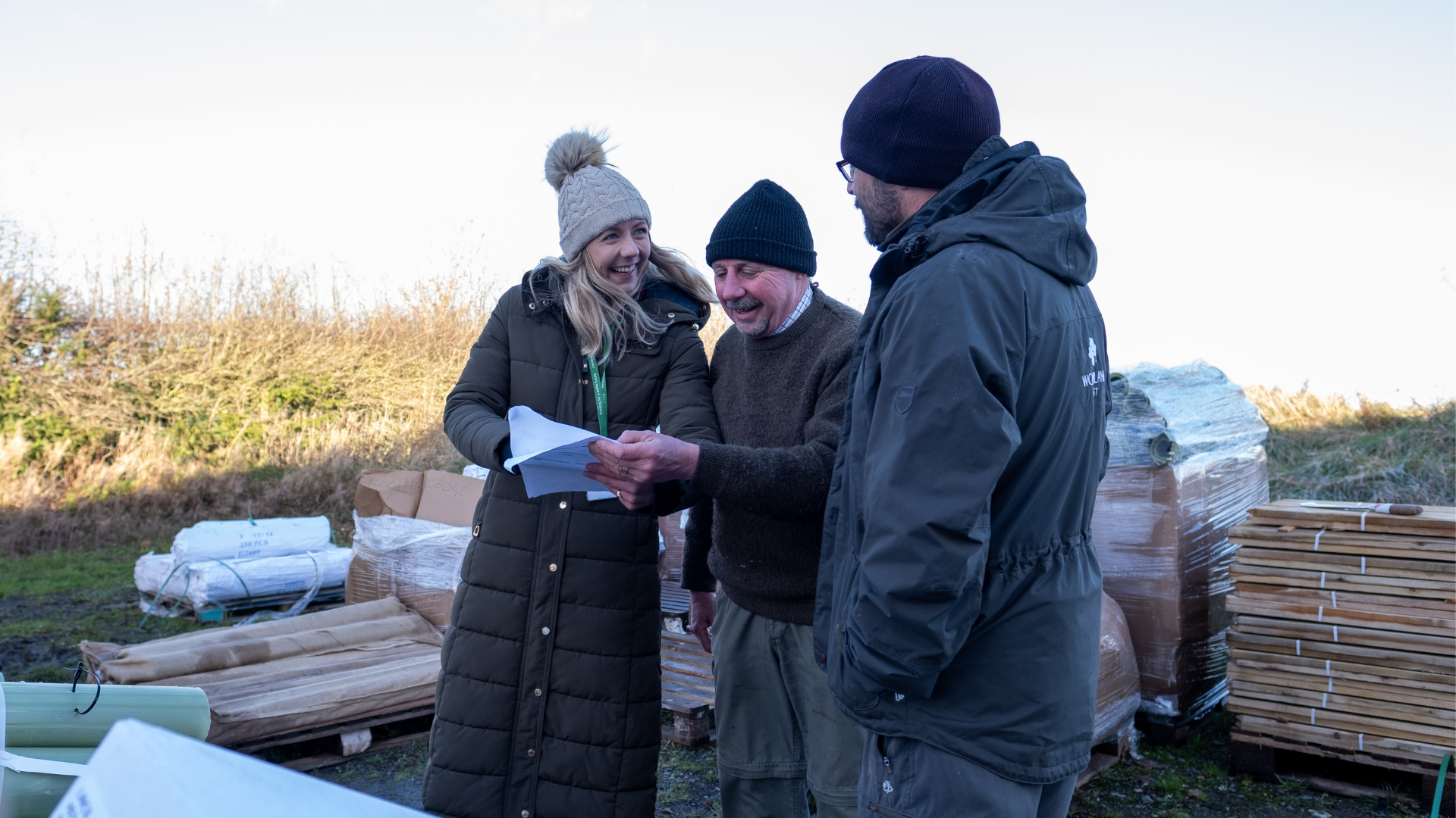 Two men and a woman look over a document, surrounded by saplings and stakes for tree planting