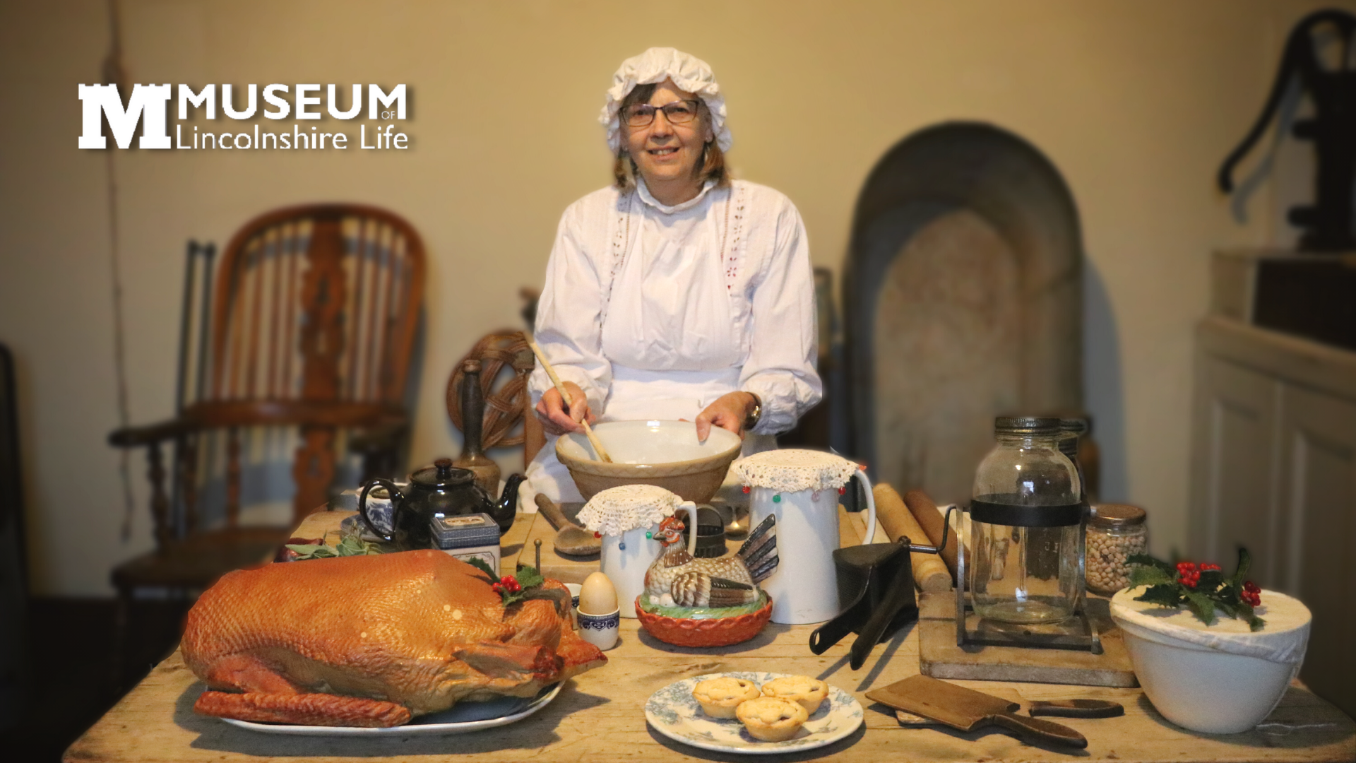 A woman dressed in a Victorian outfit in front of a table displaying Victorian food and kitchen utensils.