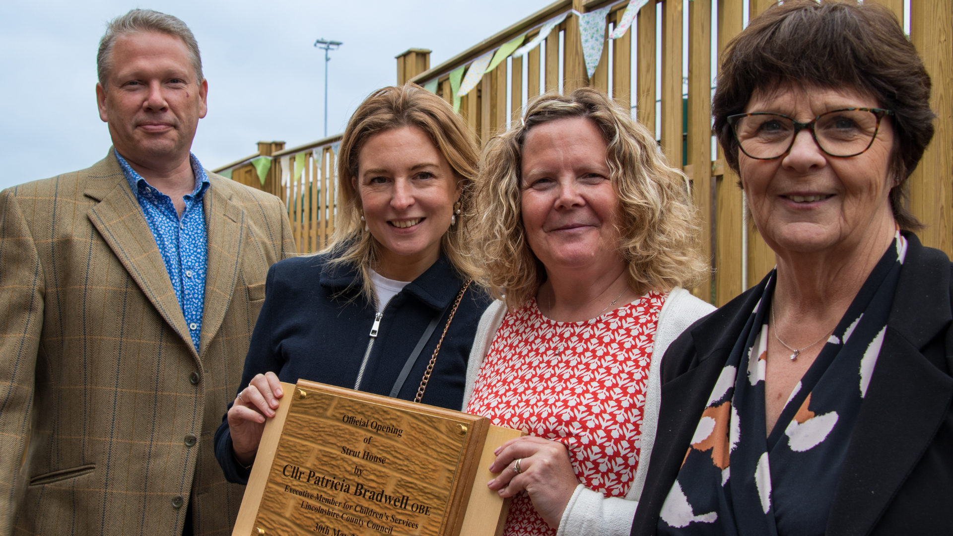 A man to the left in a suit and three women dressed smart. The second woman from the right is holding a school opening plaque in gold, proudly.