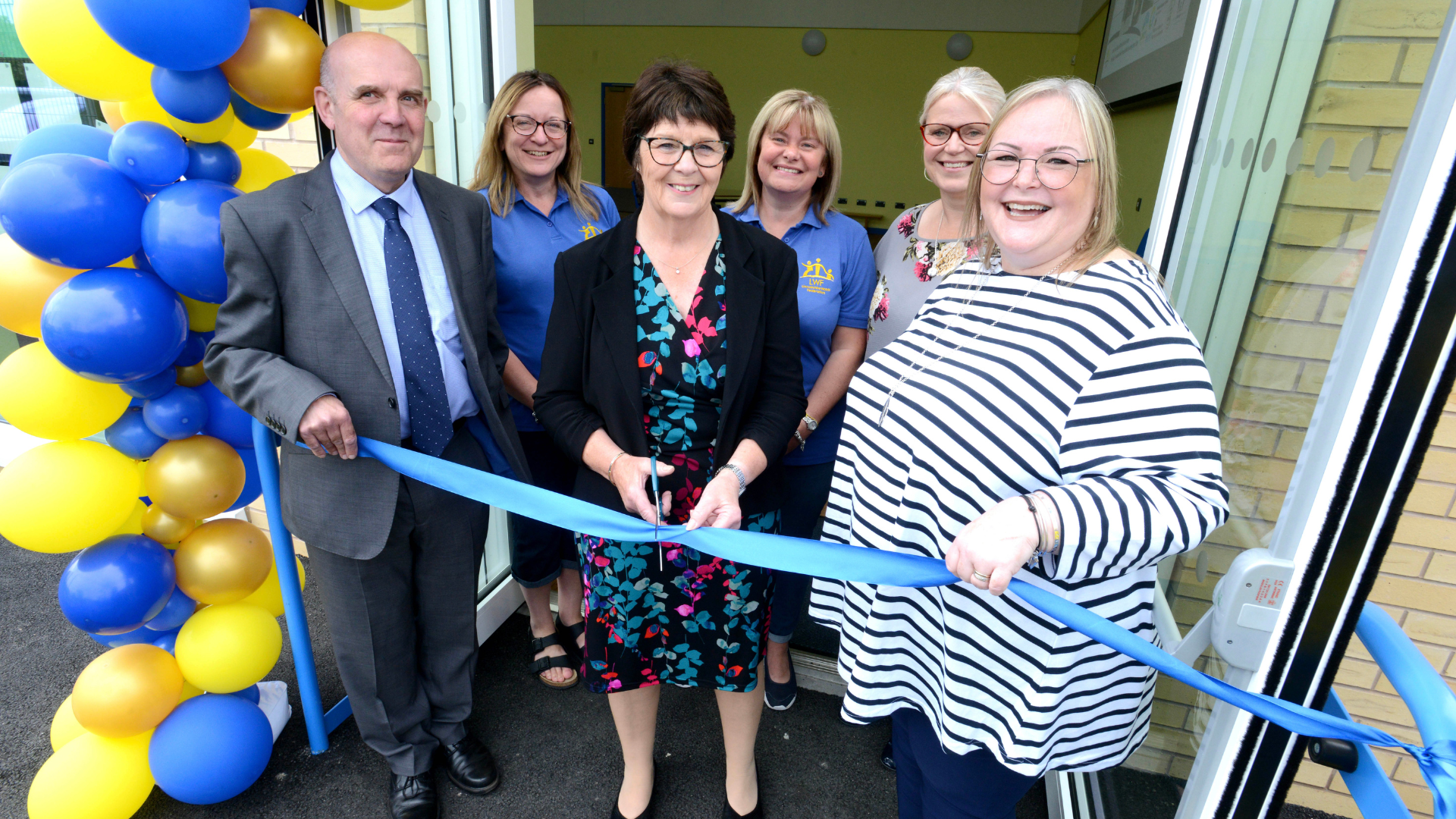 Six adults standing in smart clothes holding an opening event ribbon with blue and yellow balloons behind them