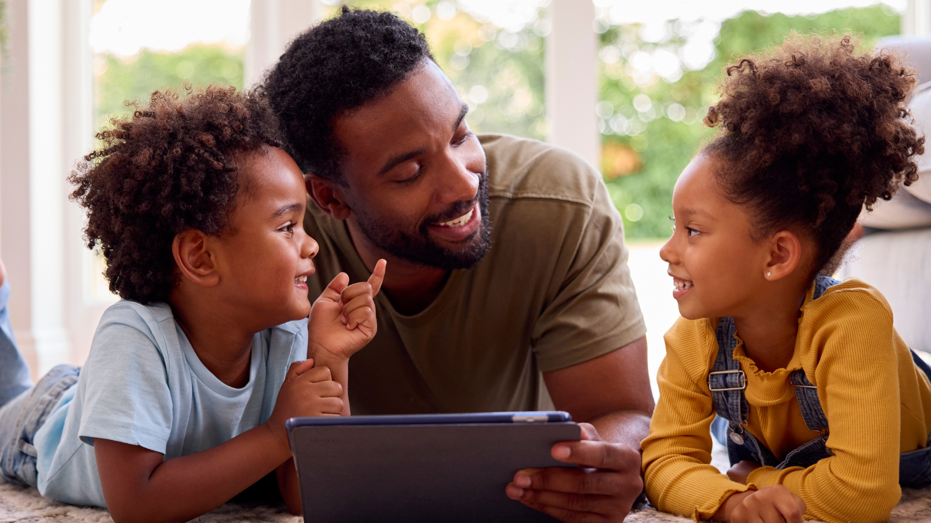 A father talking to his two young daughter holding a tablet, laying on a rug