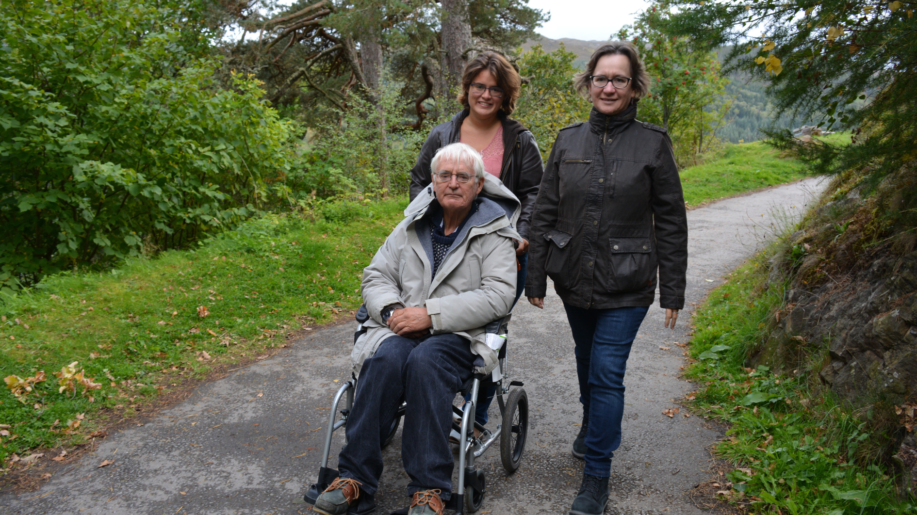 A mother and daughter walking in the countryside pushing an elderly gentleman in a wheelchair