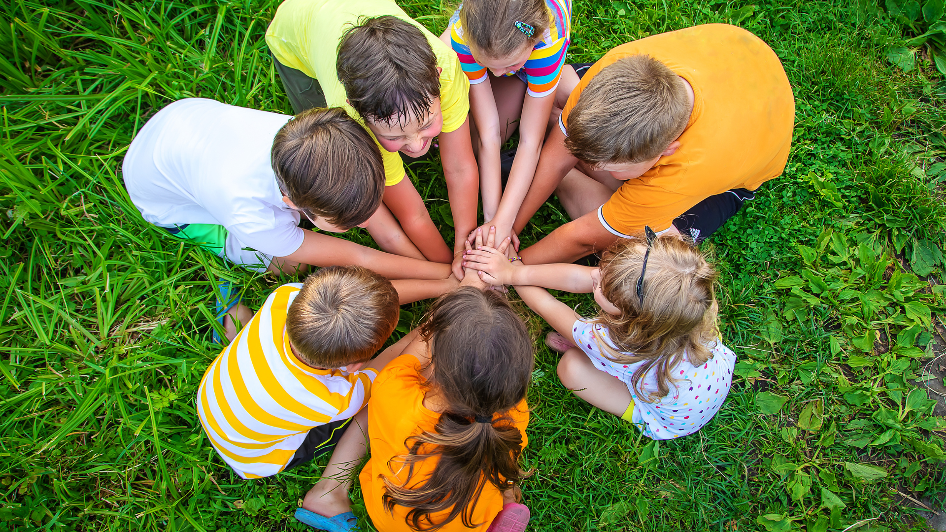 Seven children putting hands together in a circle sat on grass.