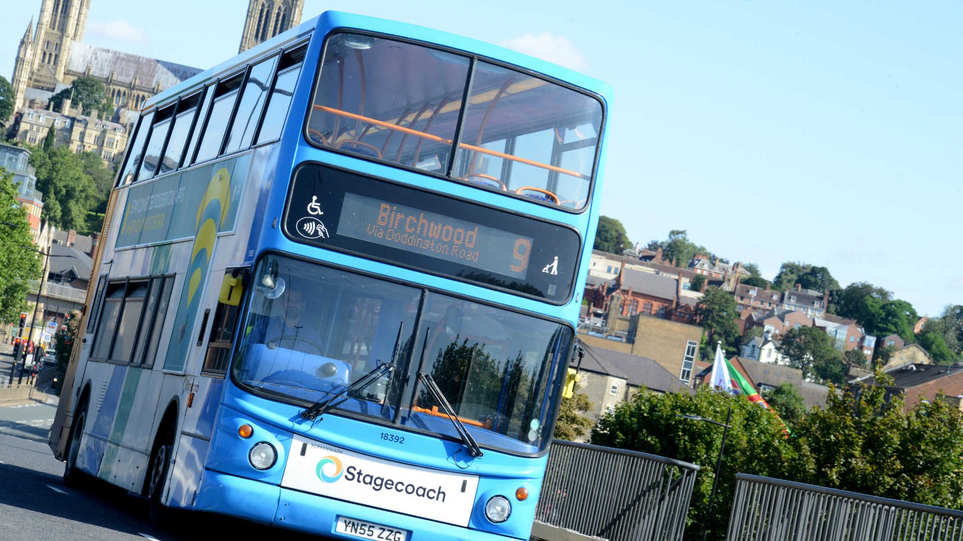 A blue double decker bus on a road with houses and trees in the background