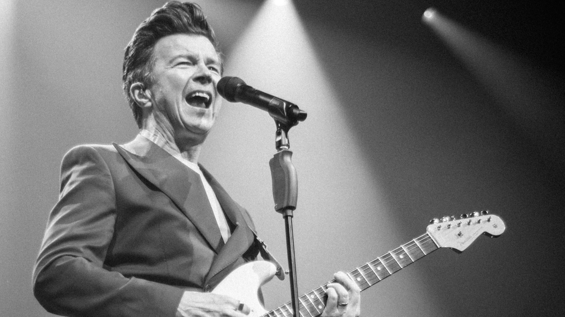 A black and white photo of Rick Astley performing on a stage and playing guitar